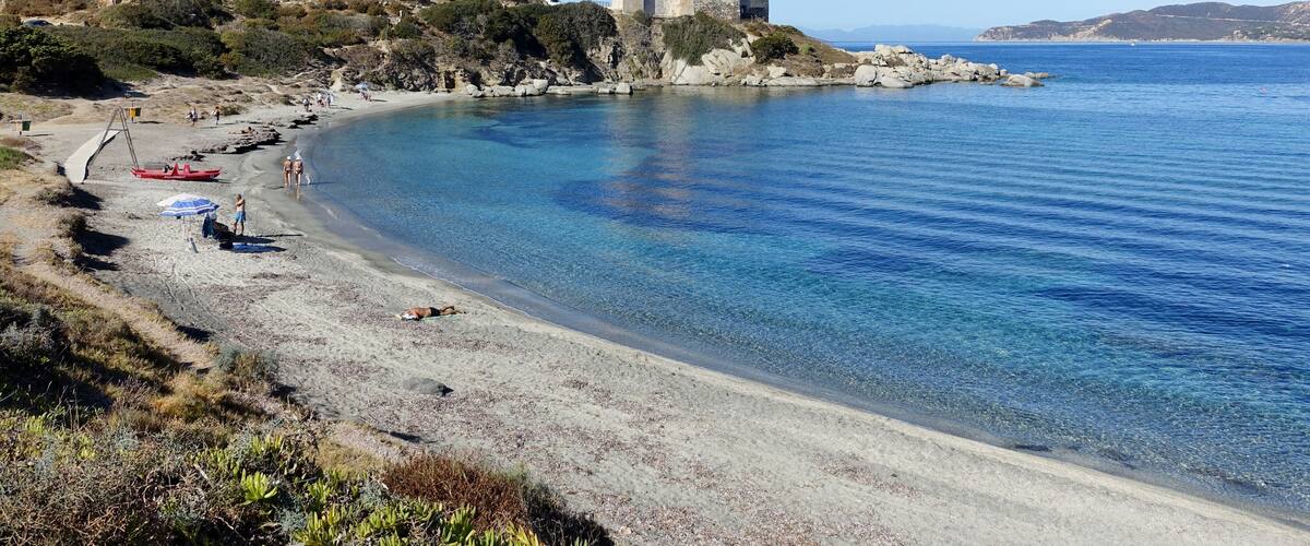 A coastal landscape with a clear blue sea, a sandy beach, rocky outcrops, and a historic stone tower on a hill.