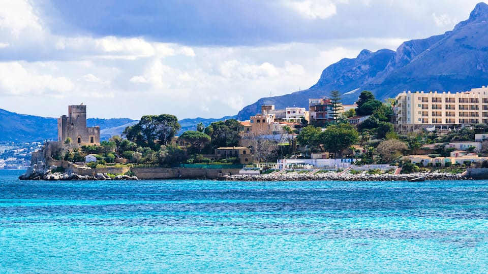A coastal town with a prominent tower, buildings, and mountains in the background.