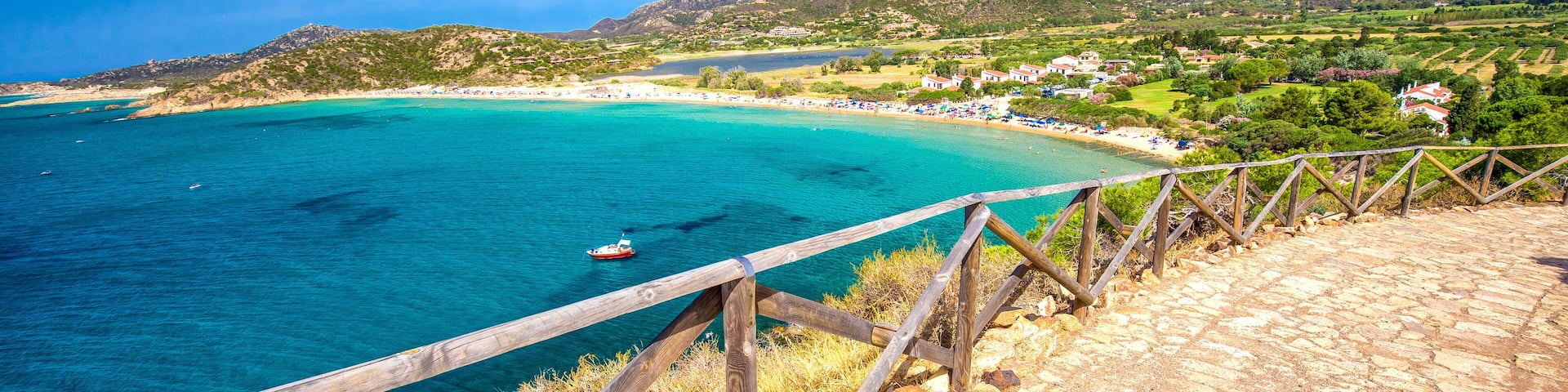 A coastal landscape with a wooden walkway overlooking turquoise waters, a boat, and a hillside village.