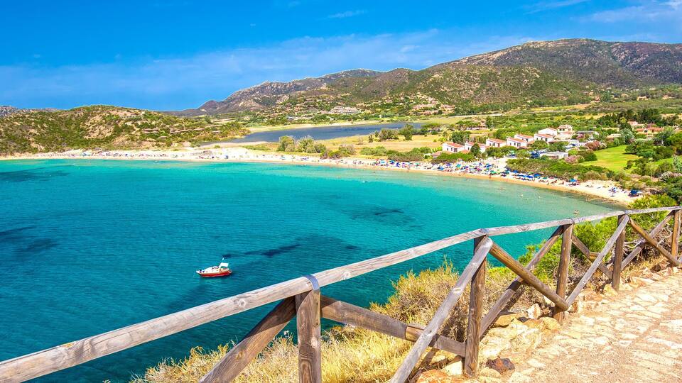 A coastal landscape with a wooden walkway overlooking turquoise waters, a boat, and a hillside village.