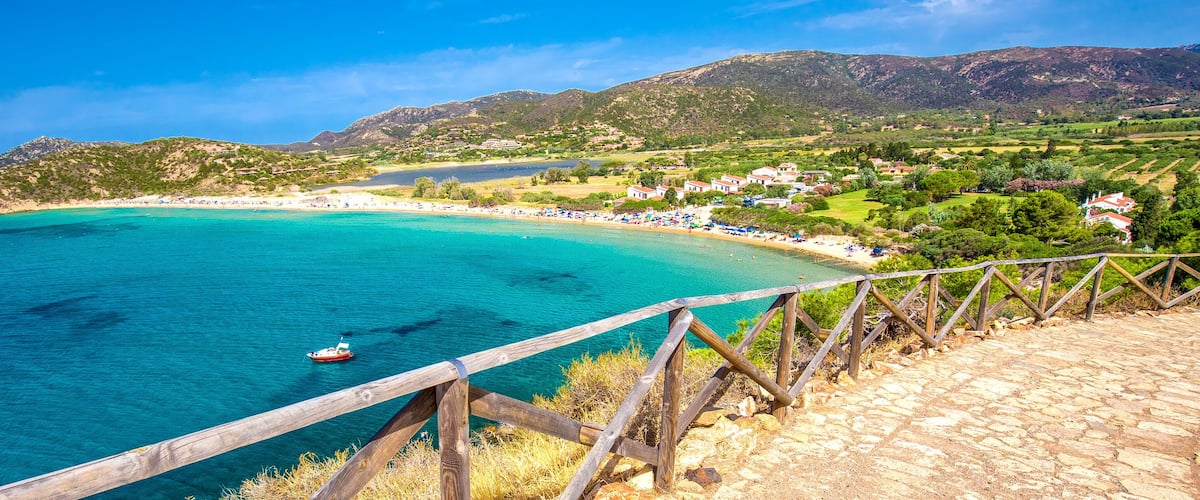 A coastal landscape with a wooden walkway overlooking turquoise waters, a boat, and a hillside village.