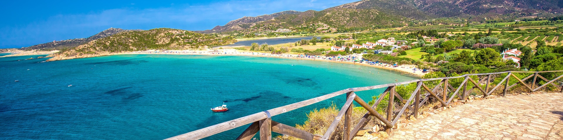 A coastal landscape with a wooden walkway overlooking turquoise waters, a boat, and a hillside village.