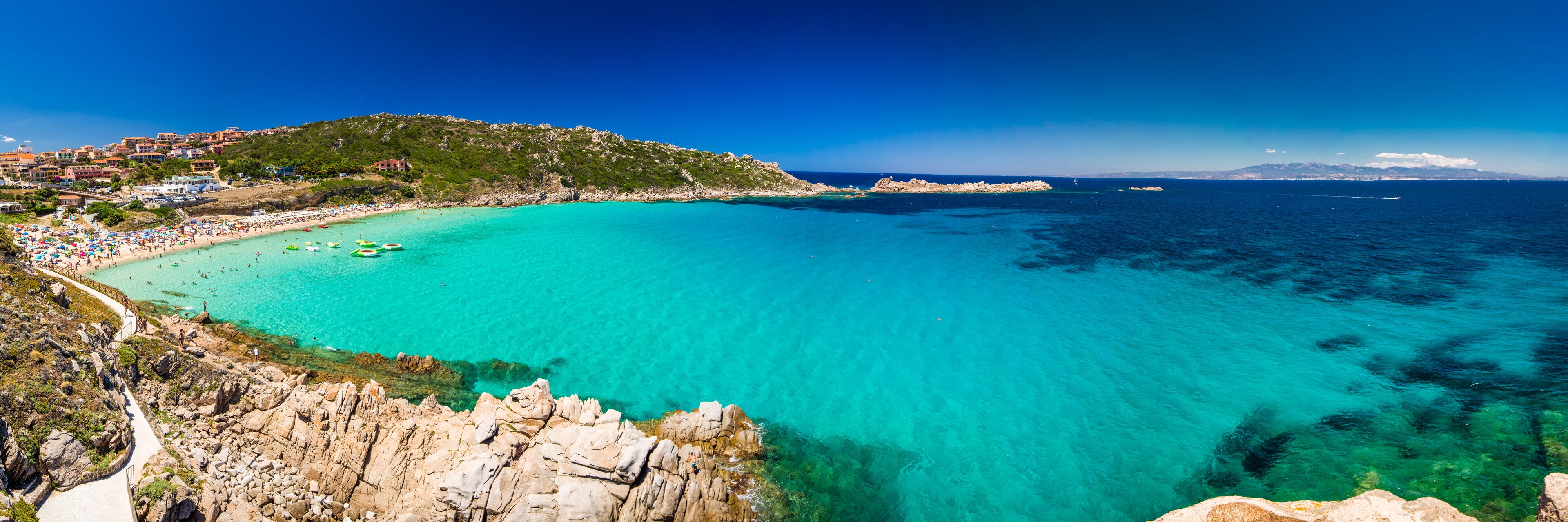 A coastal town with a turquoise bay, rocky coastline, and a hill with buildings.