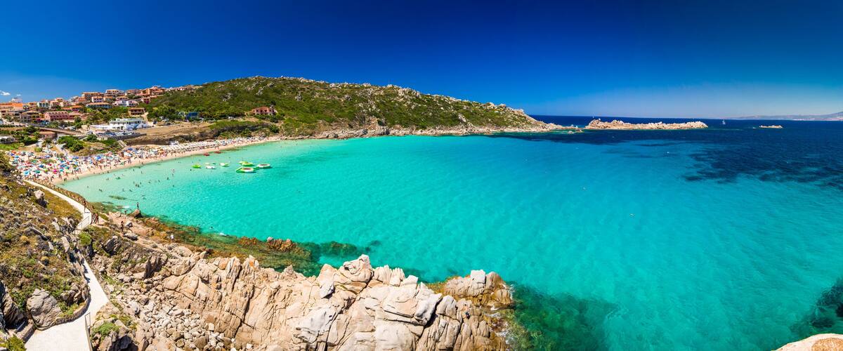 Een kustplaats met een turquoise baai, een rotsachtige kustlijn en een heuvel met gebouwen.