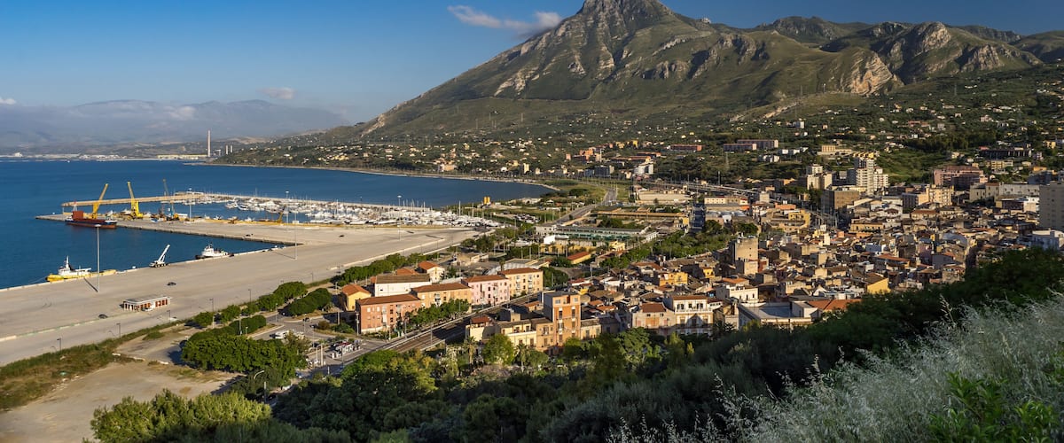 A coastal town with a marina, mountains in the background, and a mix of residential and commercial buildings.