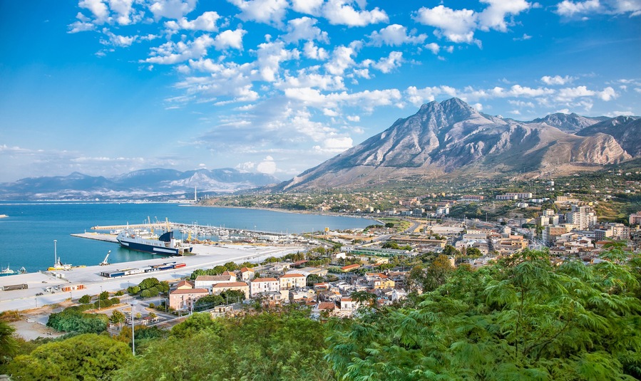 Una ciudad costera con una montaña destacada al fondo, un puerto con barcos y una combinación de paisajes urbanos y naturales.