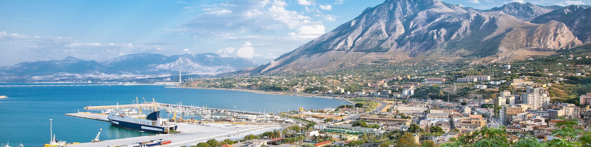 Una ciudad costera con una montaña destacada al fondo, un puerto con barcos y una combinación de paisajes urbanos y naturales.