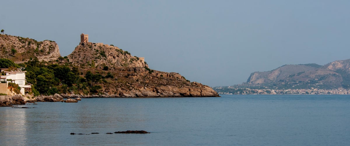 A coastal landscape with a rocky hill featuring a tower, calm sea, and distant cityscape.