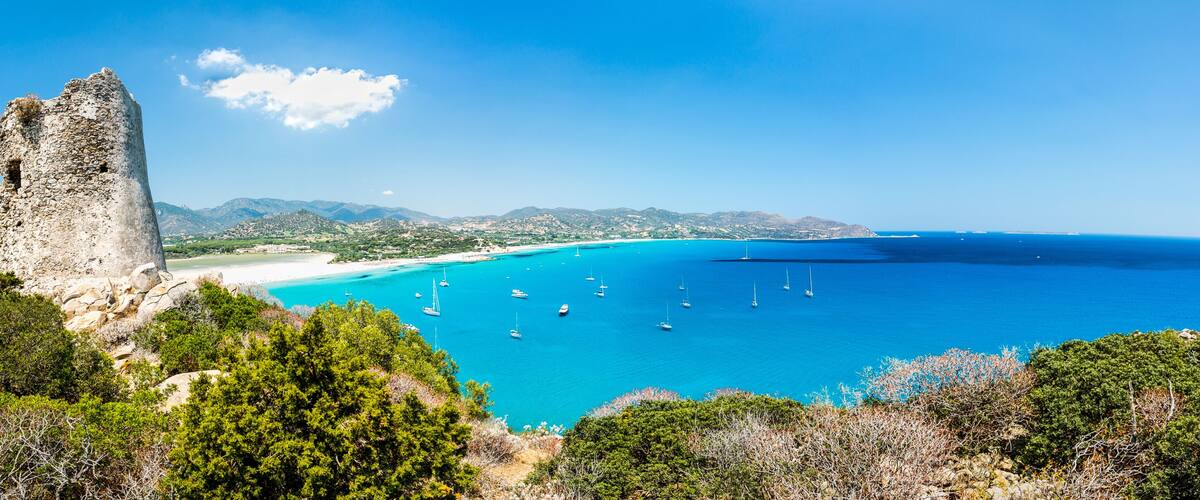 A coastal landscape with a historic stone tower, clear turquoise waters, and a sandy beach.