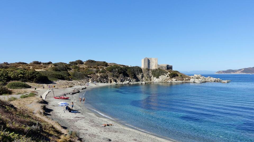 A coastal landscape with a clear blue sea, a sandy beach, and a historic stone tower on a rocky outcrop.