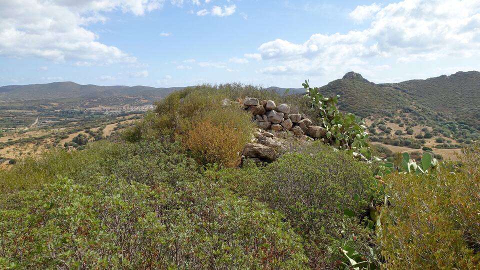 A mountainous landscape with a stone wall, green shrubs, and a clear sky.