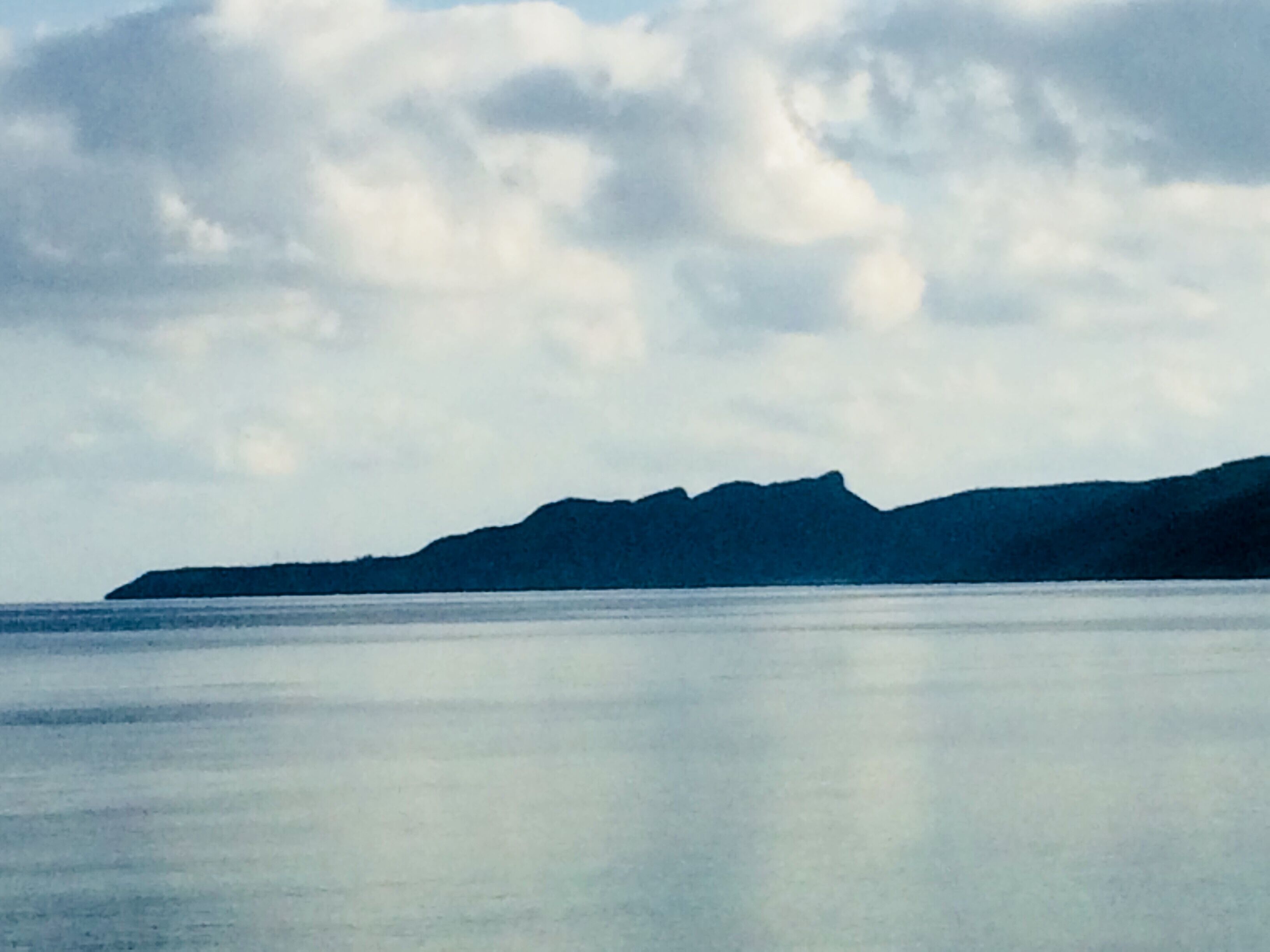 A calm sea with a mountain range in the distance under a cloudy sky.