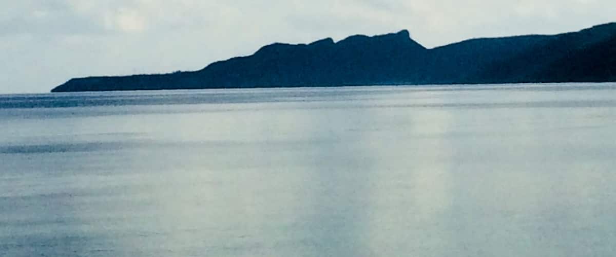 A calm sea with a mountain range in the distance under a cloudy sky.