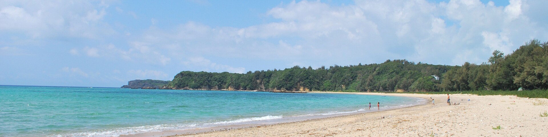 A sandy beach with clear turquoise water and a forested hillside in the background.
