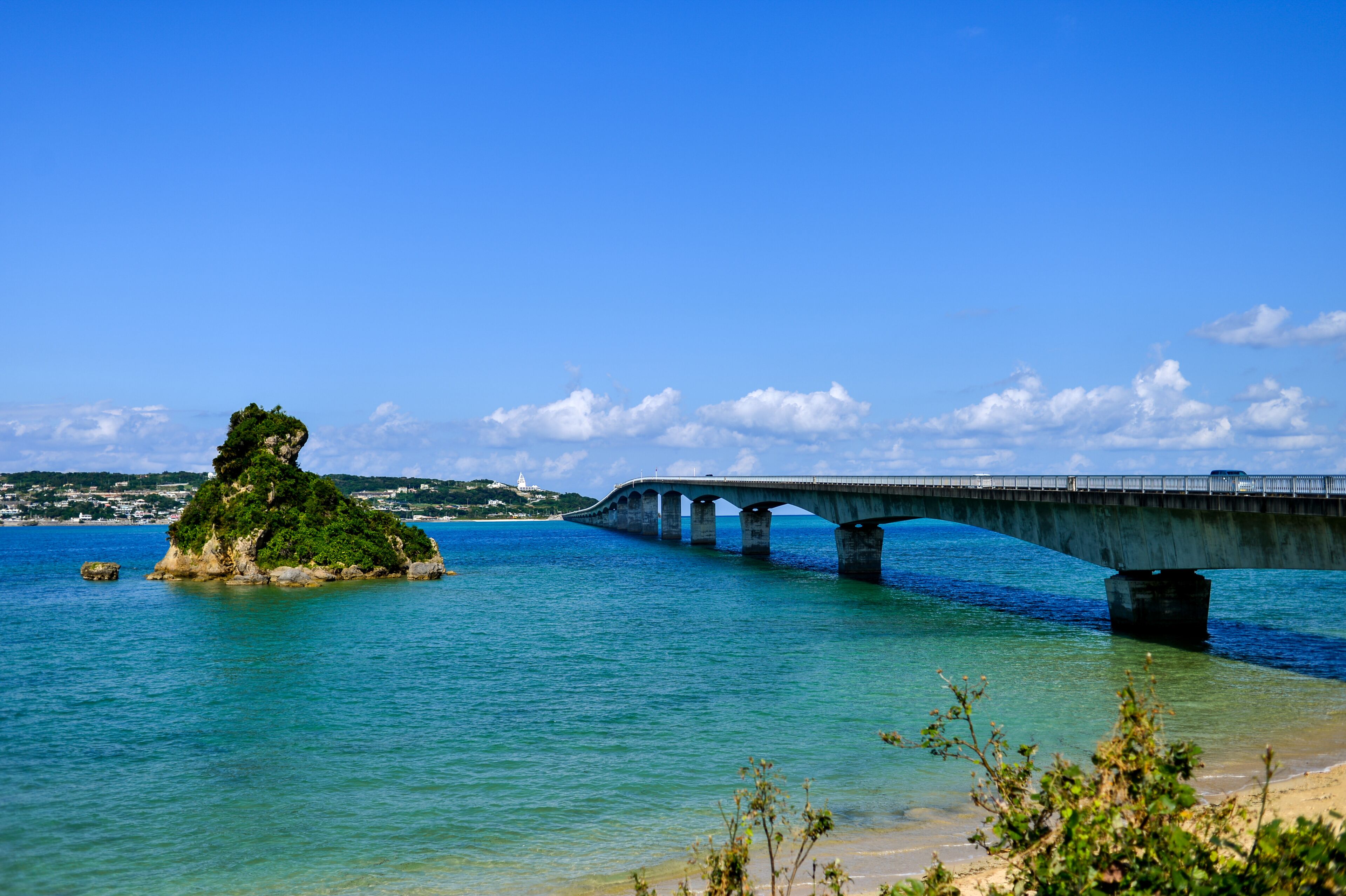 Uma ponte sobre águas turquesa, uma ilha verde e um céu azul límpido.