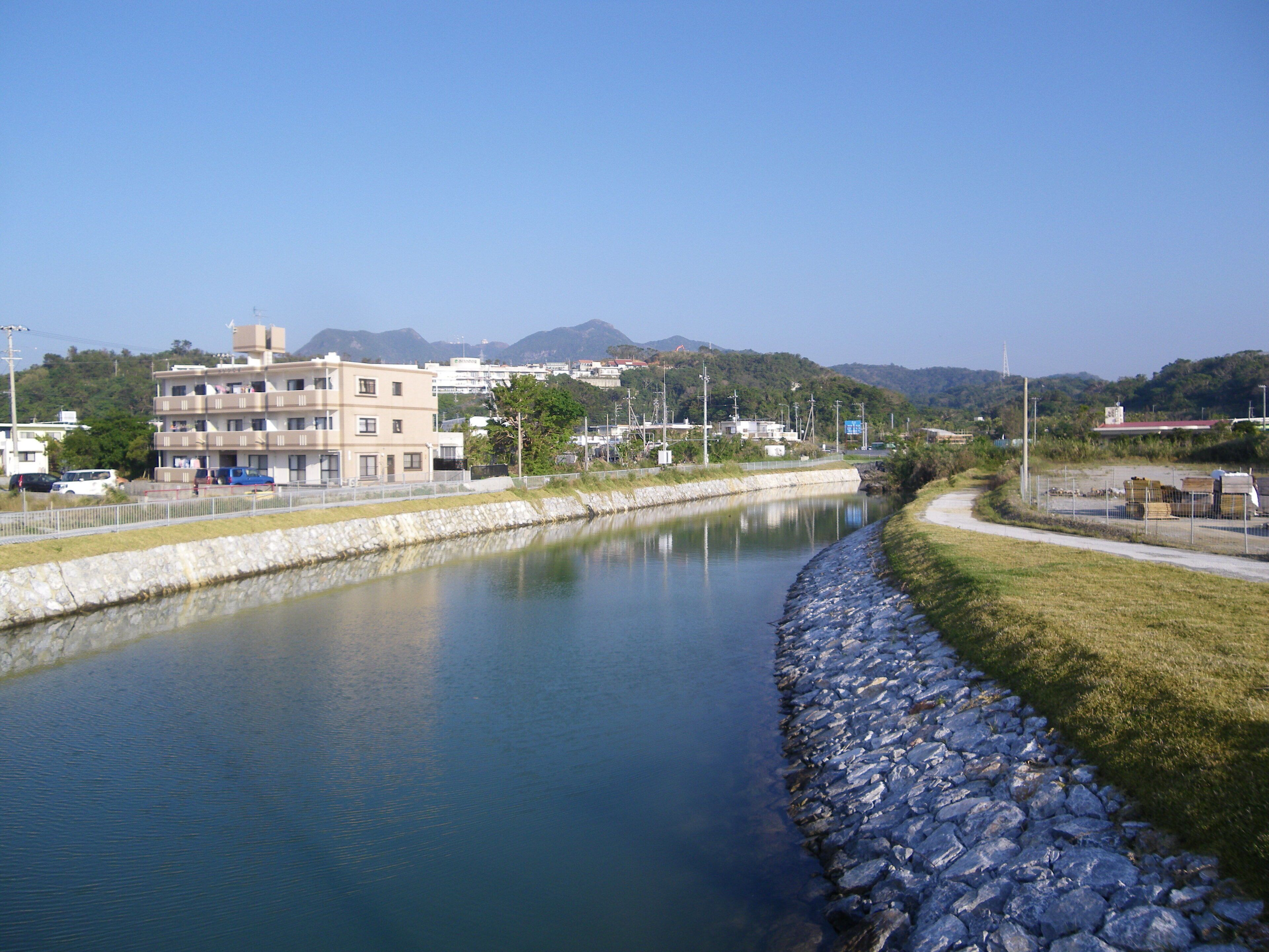 A calm river with a stone embankment, buildings, and mountains in the background.