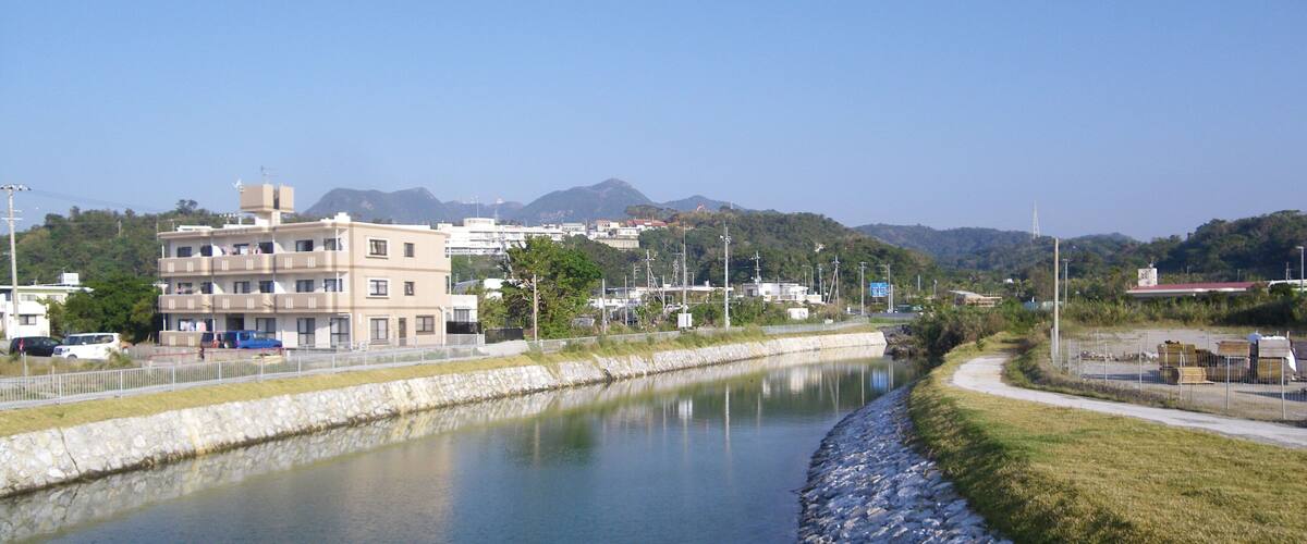 A calm river with a stone embankment, buildings, and mountains in the background.