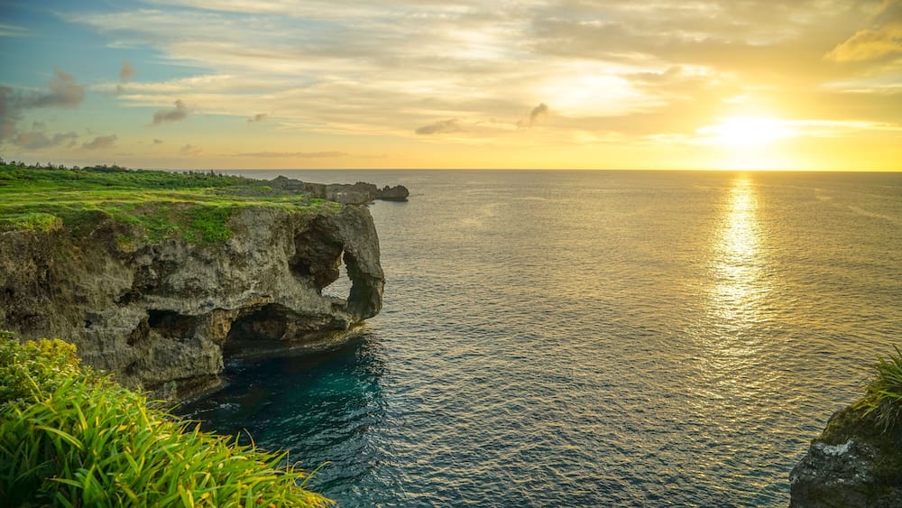 A coastal landscape with a prominent rock formation featuring a natural arch, lush greenery, and a sunset over the ocean.