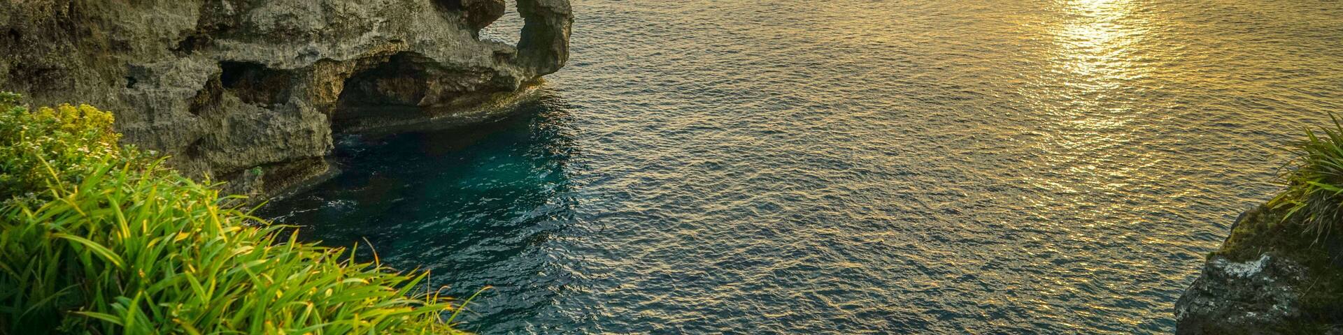 A coastal landscape with a prominent rock formation featuring a natural arch, lush greenery, and a sunset over the ocean.