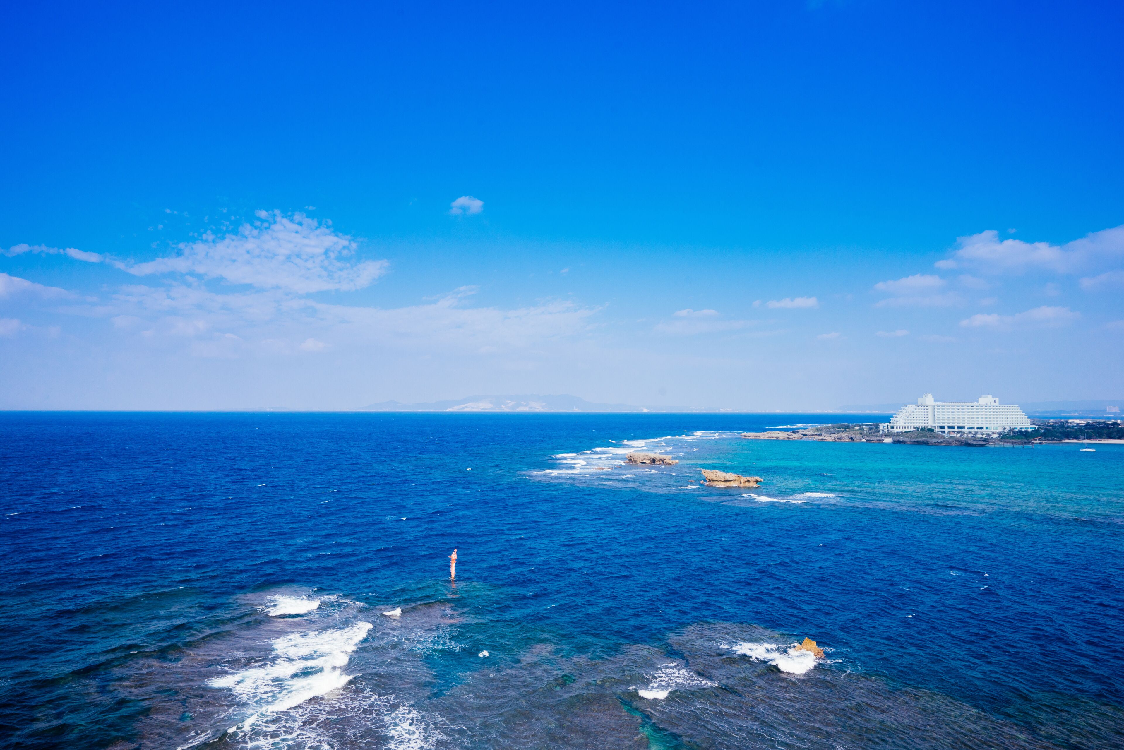Uma cena costeira com céu azul límpido, ondas do oceano, um farol e um edifício branco com varanda.