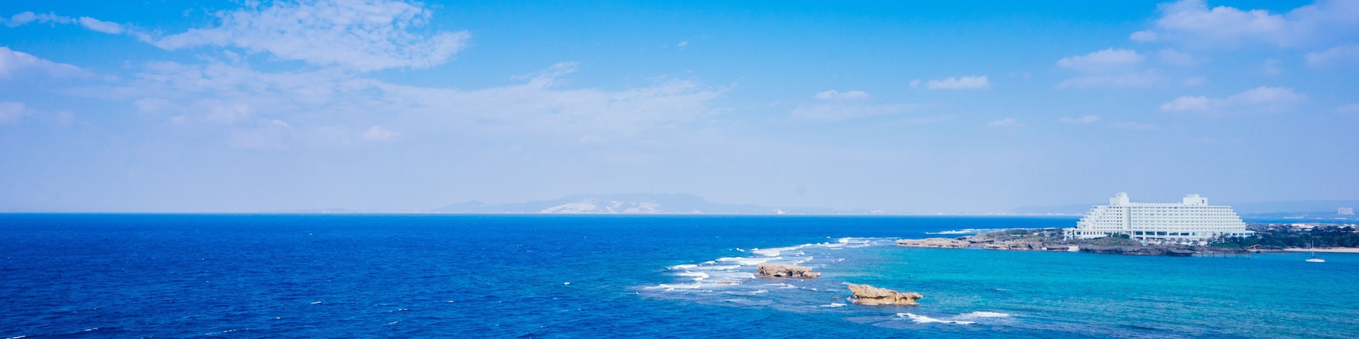 Uma cena costeira com céu azul límpido, ondas do oceano, um farol e um edifício branco com varanda.