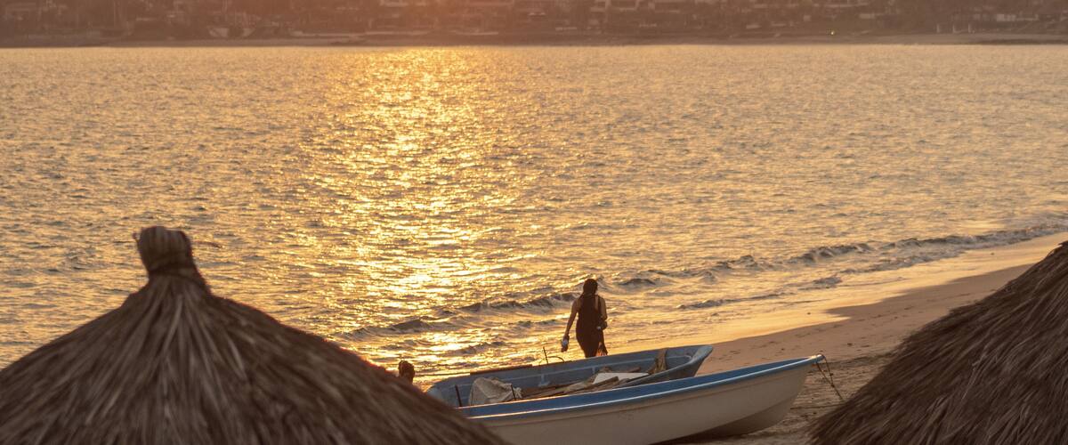 A beach with a boat and thatched huts at sunset.
