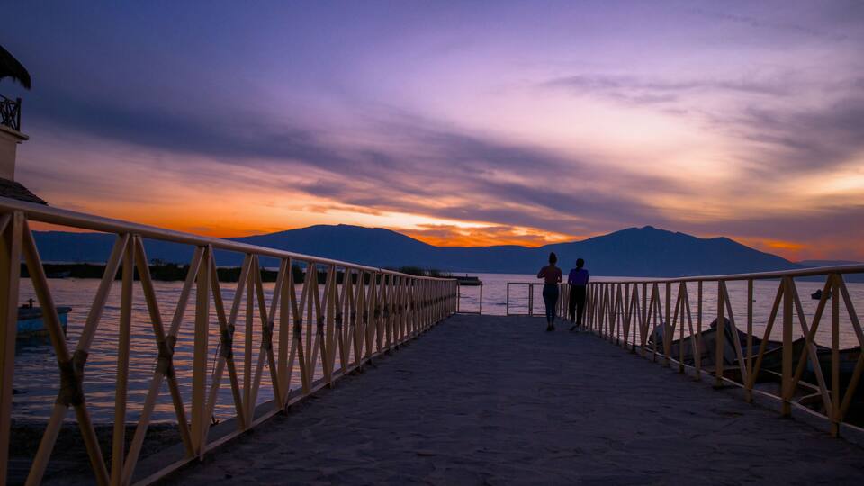 A sunset over a lake with mountains in the background and a wooden pier leading to a dock.
