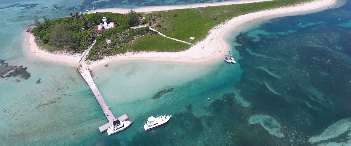 An island with a lighthouse, a white sandy beach, clear turquoise waters, and a pier.