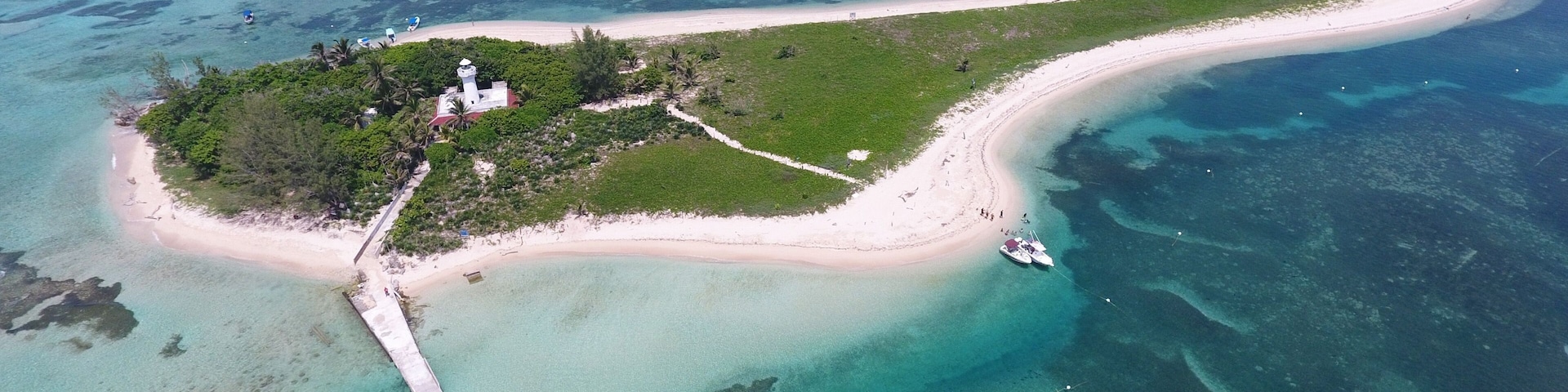 An island with a lighthouse, a white sandy beach, clear turquoise waters, and a pier.