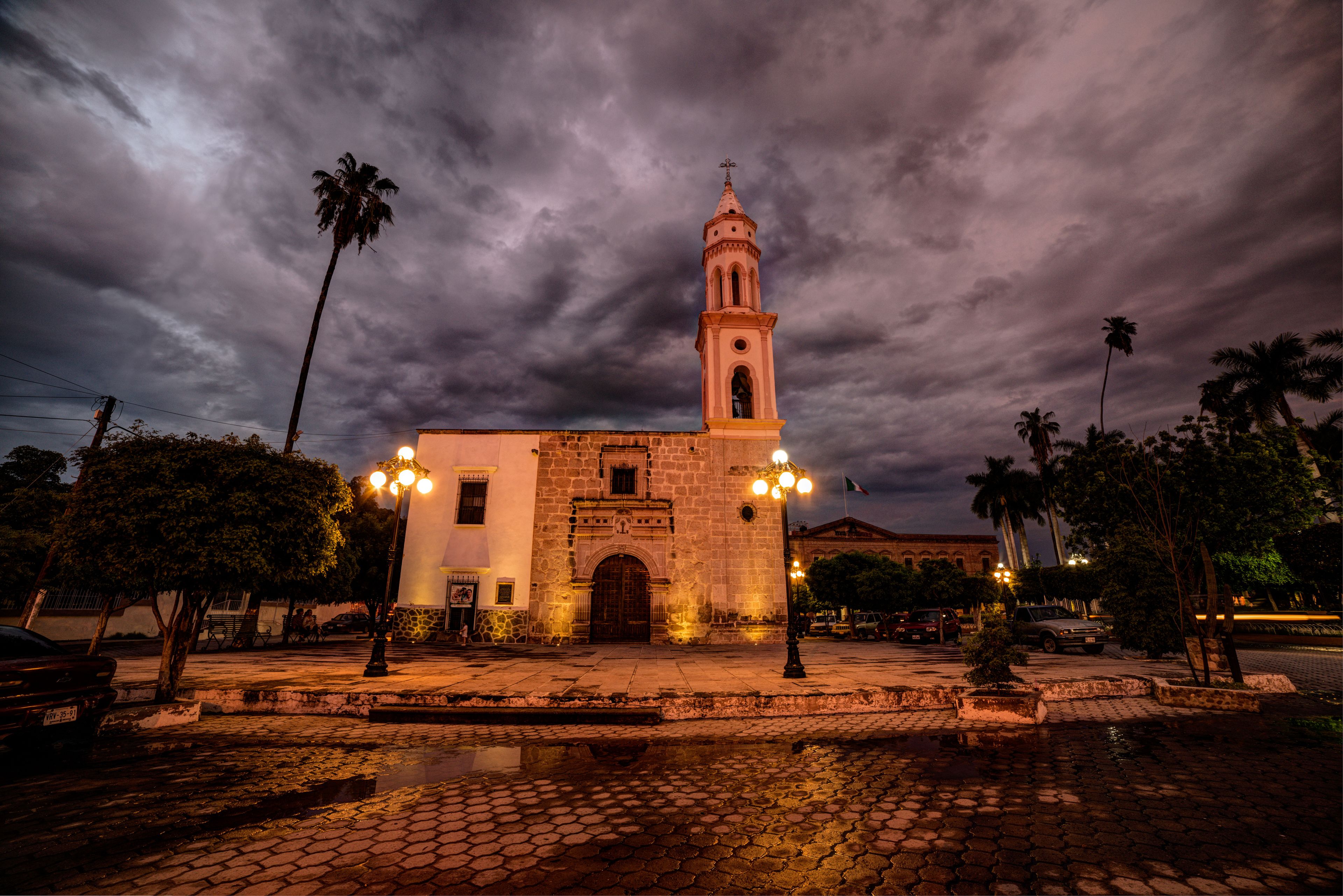 A historic building with a bell tower, surrounded by palm trees and cobblestone streets at night.
