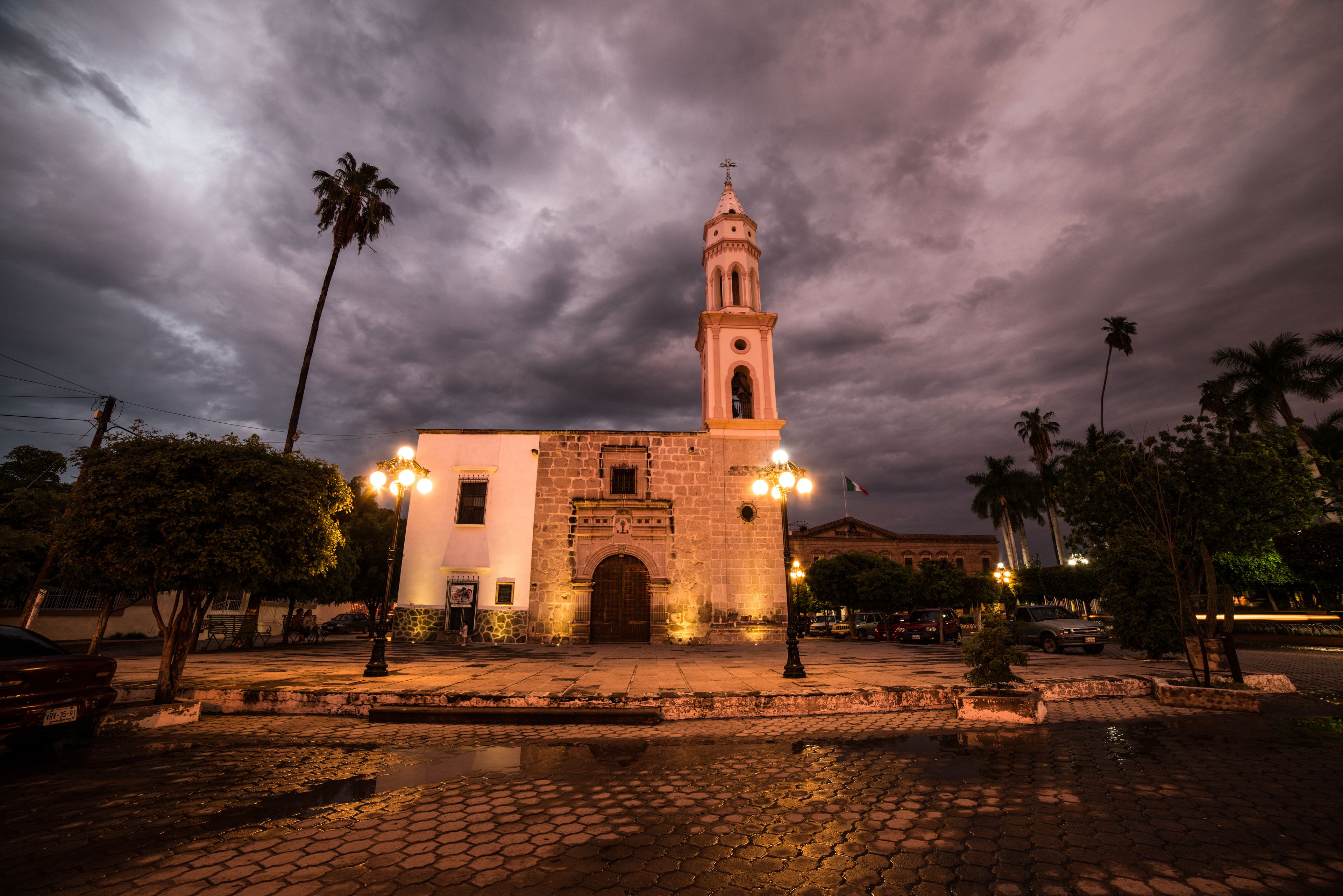 A historic building with a bell tower, surrounded by palm trees and cobblestone streets at night.