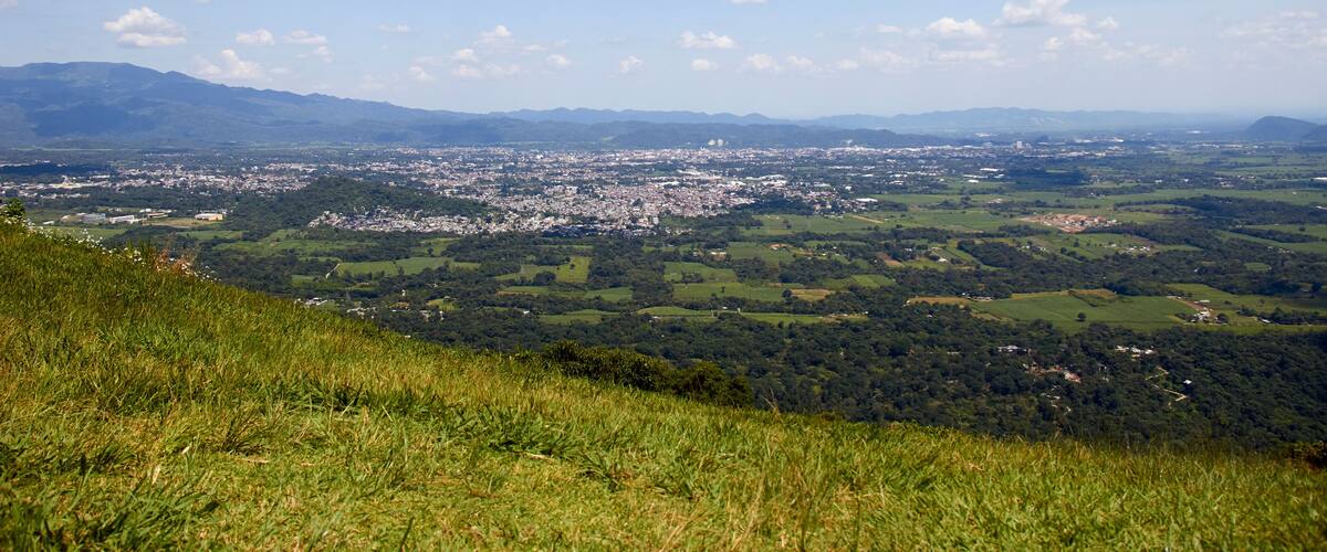 A panoramic view of a city surrounded by green hills and mountains under a partly cloudy sky.