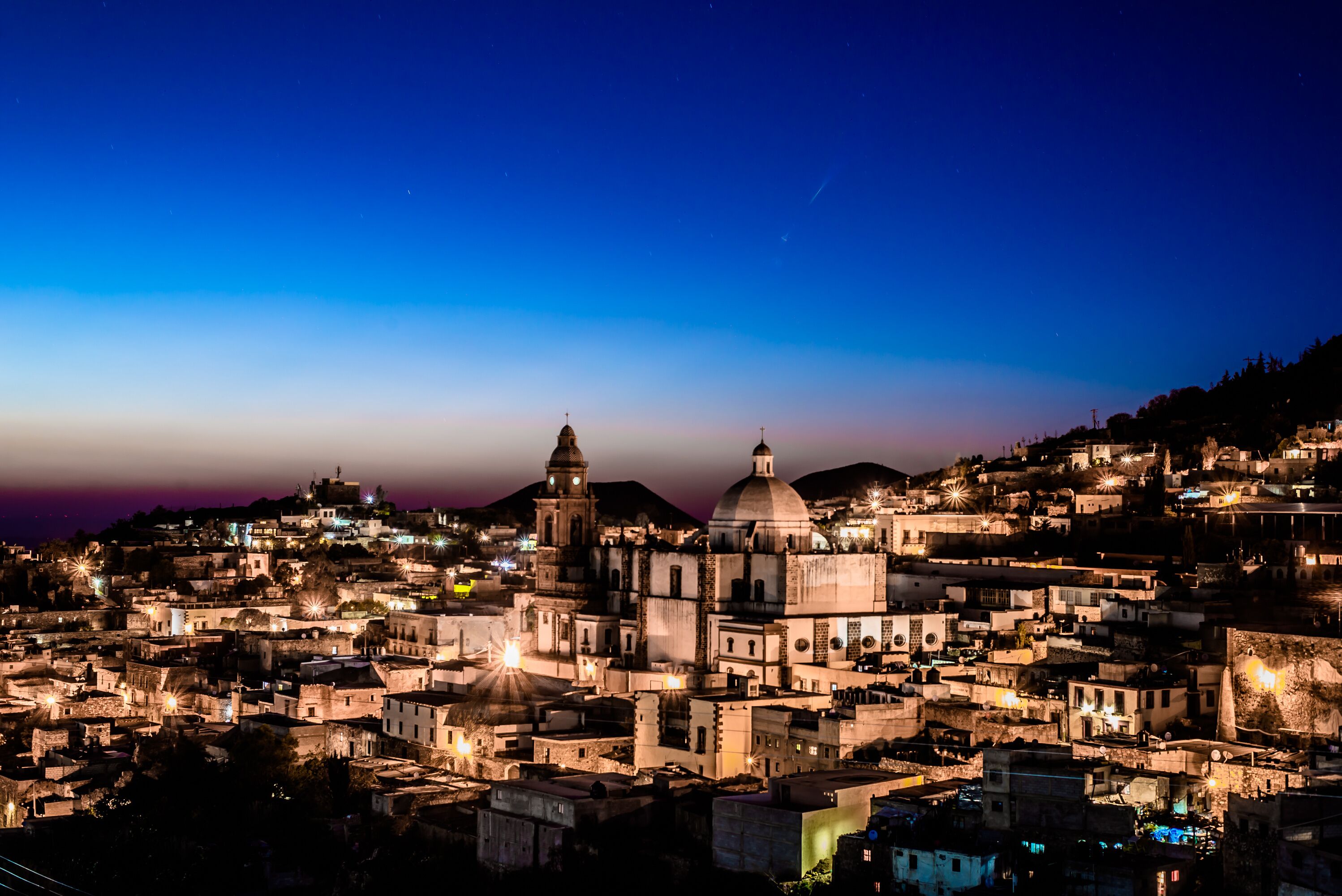 A cityscape at dusk with illuminated buildings and prominent domed structures.