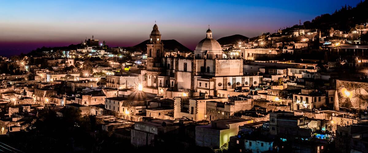 A cityscape at dusk with illuminated buildings and prominent domed structures.