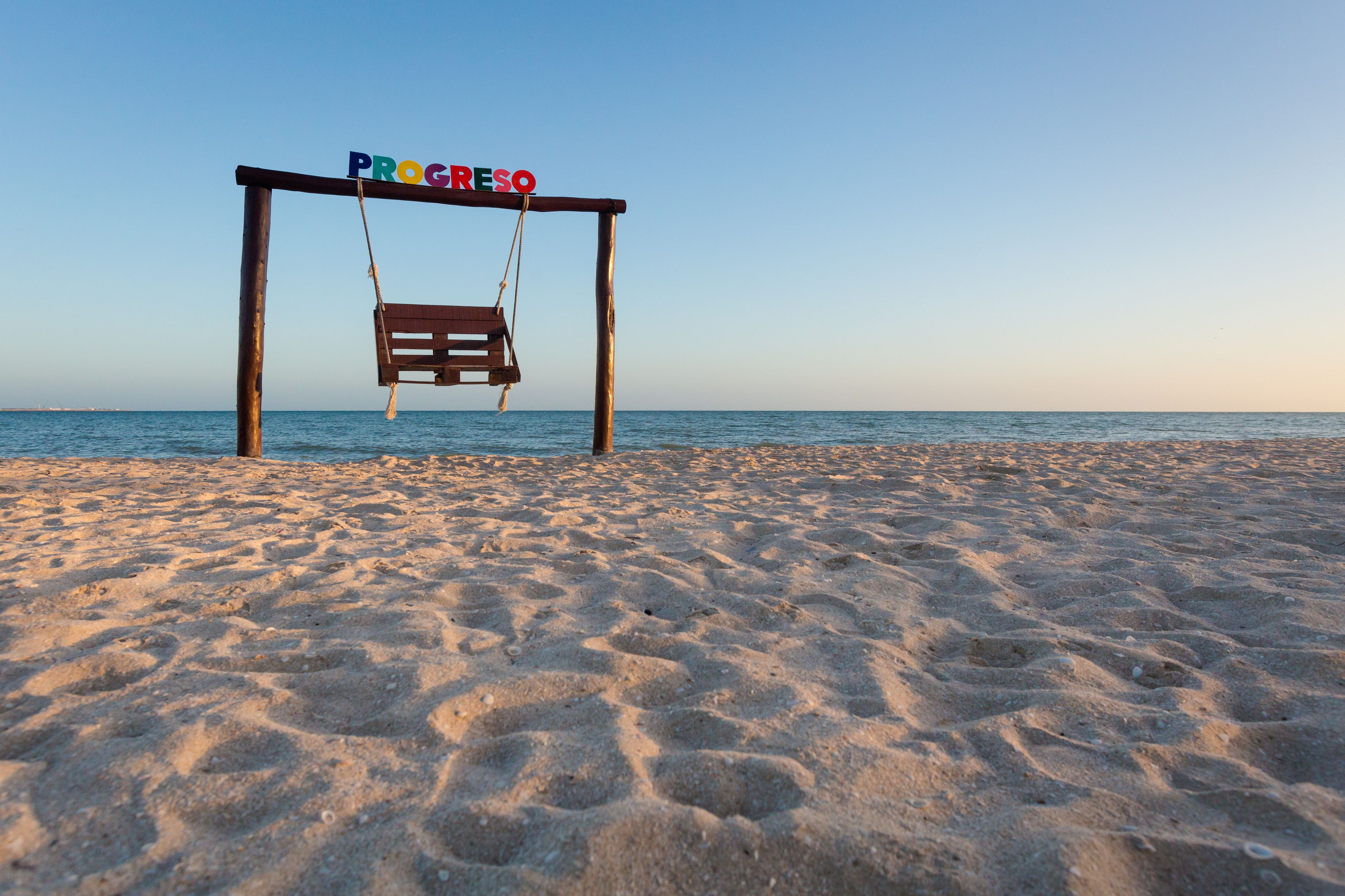 A wooden swing set with the word 'PROGRESO' on it, set on a sandy beach with the ocean in the background.