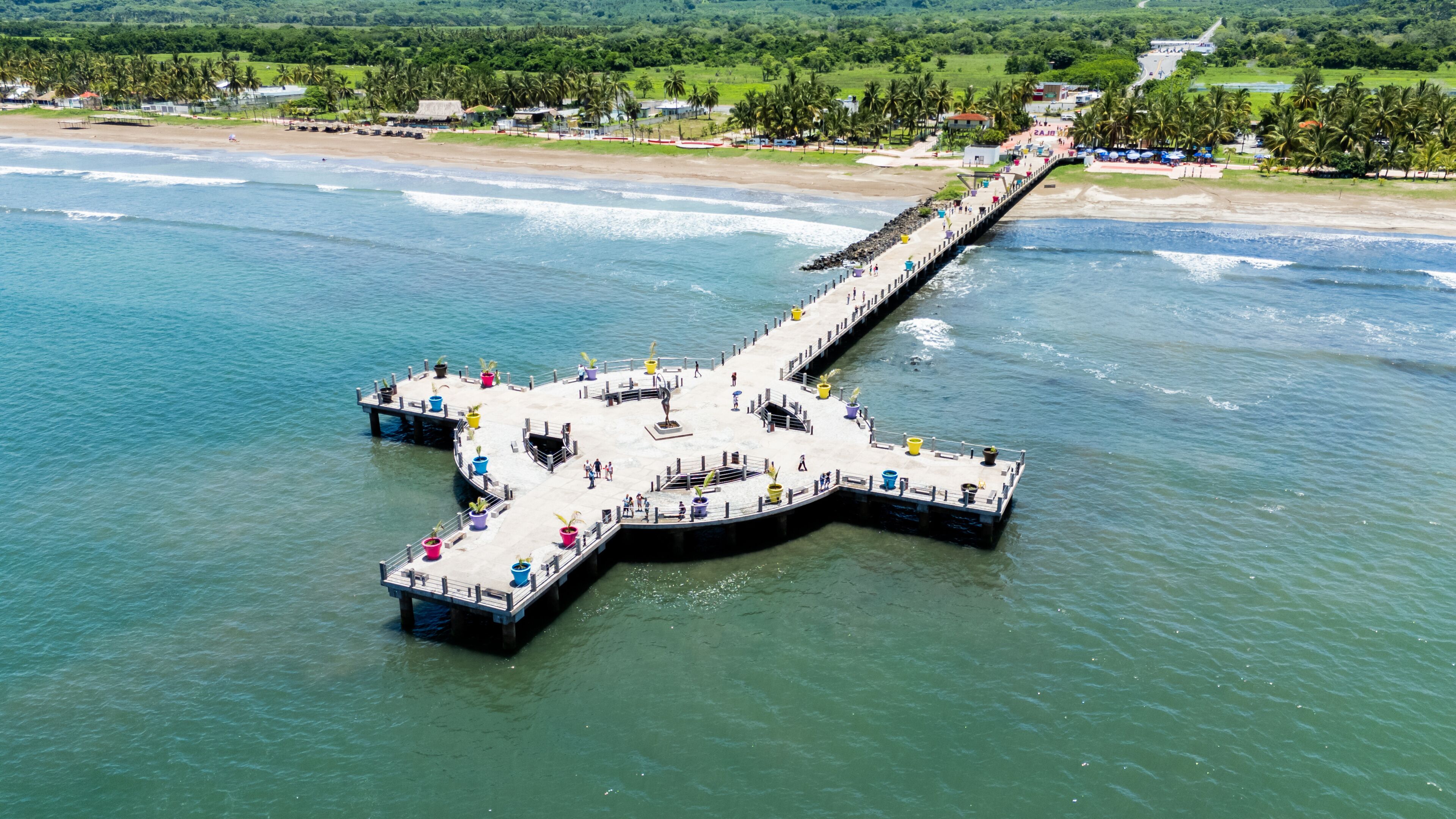 A pier extending into a body of water with a beach and greenery in the background.