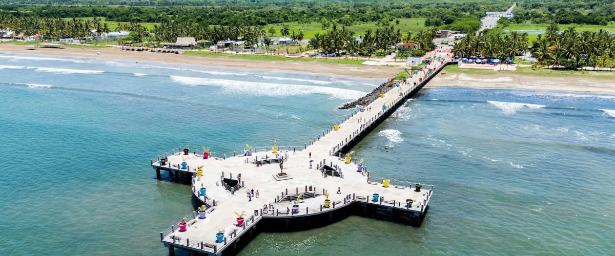 A pier extending into a body of water with a beach and greenery in the background.