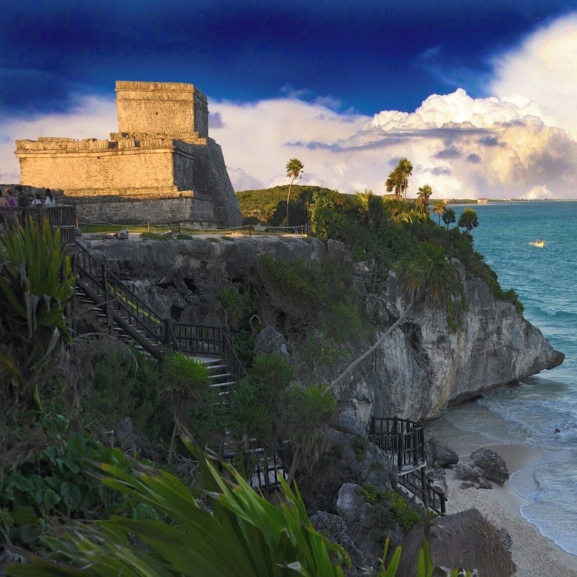 A coastal cliff with a stone structure, palm trees, and a staircase leading down to the beach.