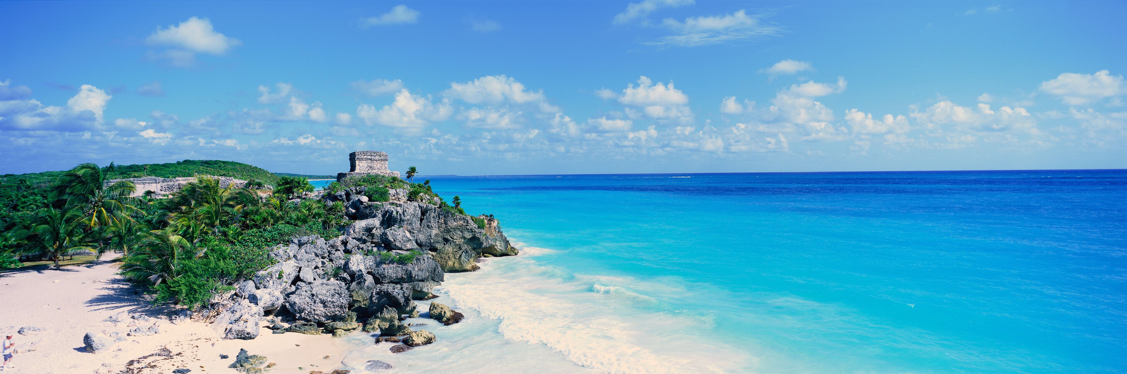 A beach with clear turquoise waters, a rocky cliff with ruins, and lush greenery.