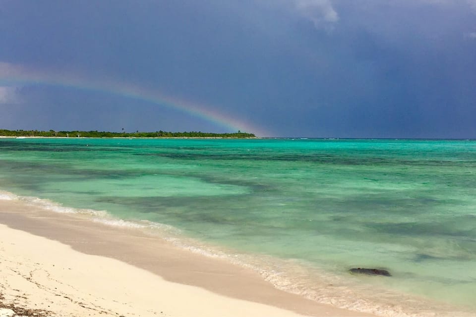 A beach with clear turquoise water, a sandy shore, and a rainbow in the sky.