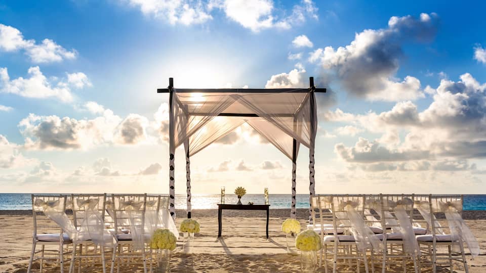 A beach ceremony setup with a canopy, white chairs, and yellow flowers.