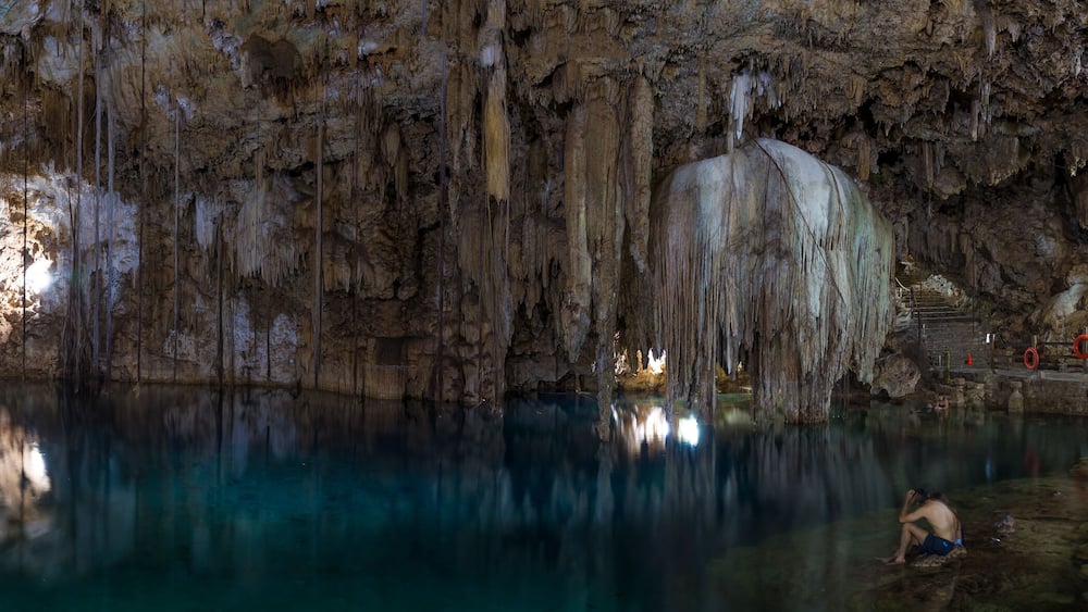 A cave with stalactites and stalagmites, a pool of water, and a person climbing a rock formation.