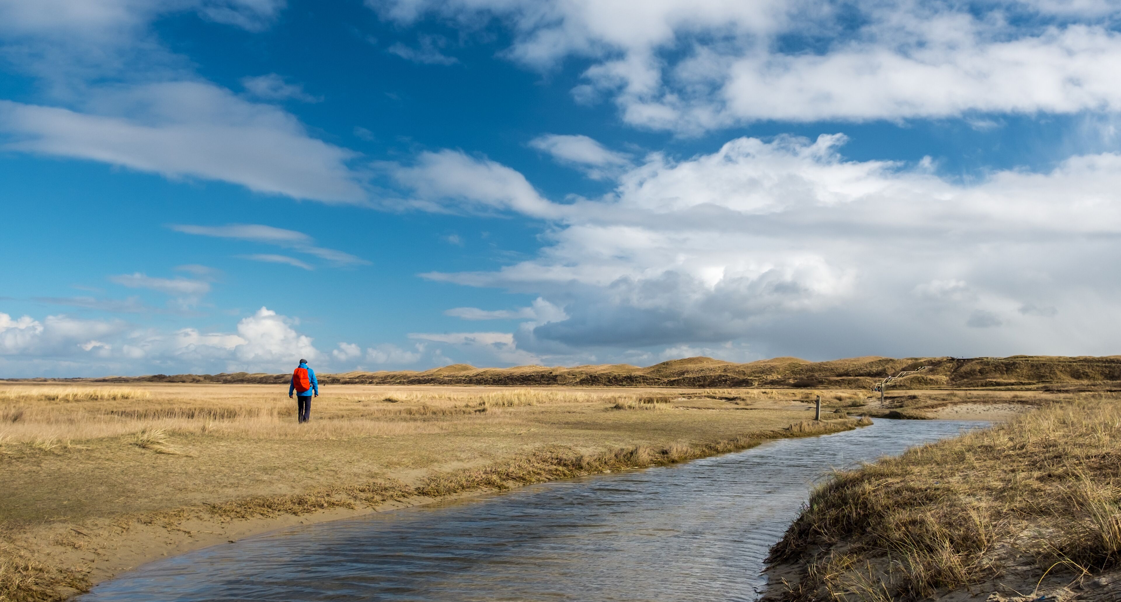 A person walking along a riverbank in a flat, grassy landscape with distant hills under a partly cloudy sky.