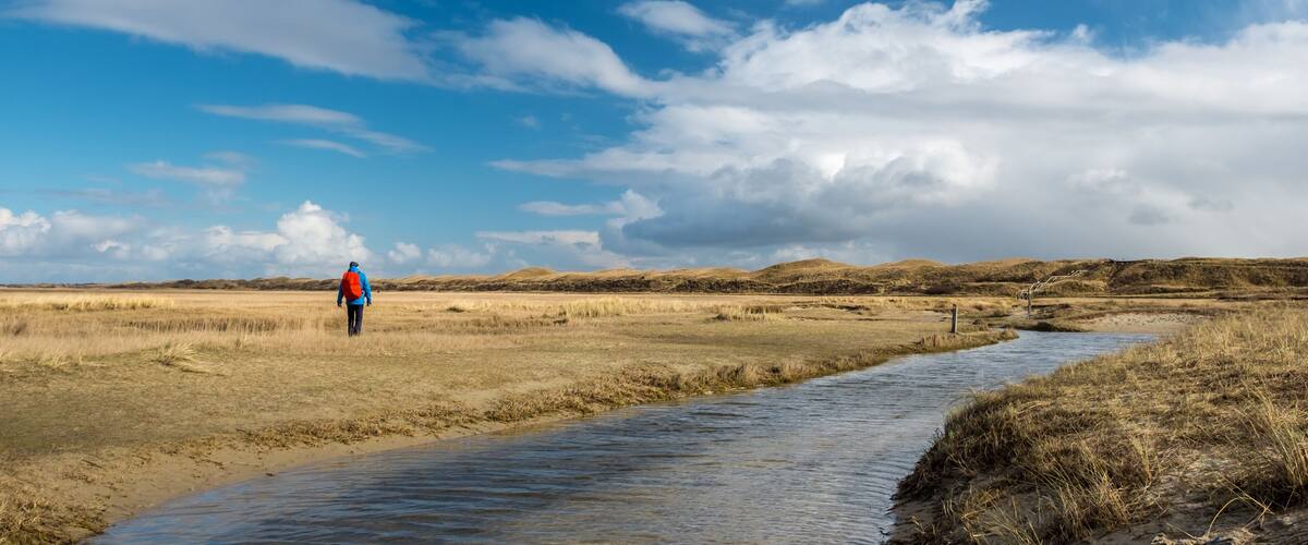 A person walking along a riverbank in a flat, grassy landscape with distant hills under a partly cloudy sky.