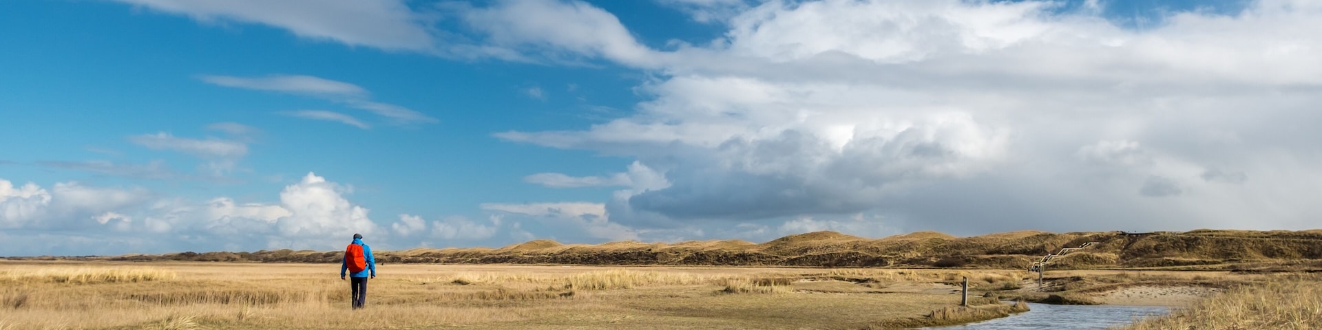 A person walking along a riverbank in a flat, grassy landscape with distant hills under a partly cloudy sky.