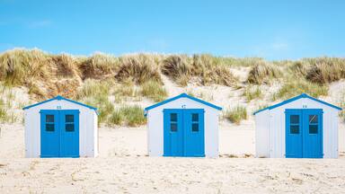 Three beach huts with blue doors and white walls, numbered 16 and 17, on a sandy beach with dunes in the background.