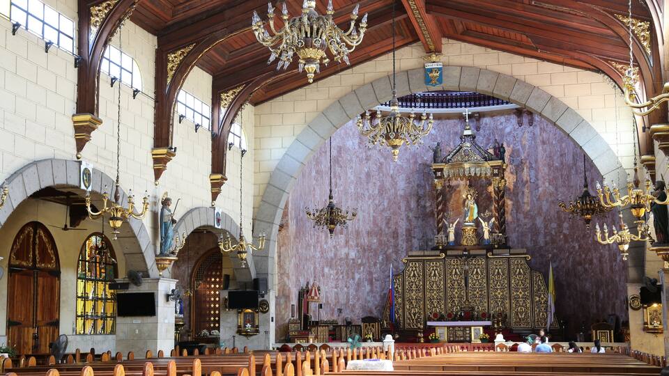 An ornate church interior with high wooden ceilings, chandeliers, and a central altar.