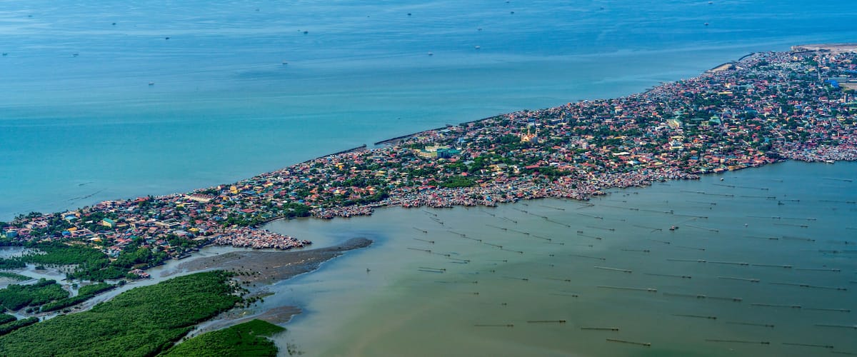 An aerial view of a coastal city with dense urban development along the shoreline, a river delta, and scattered boats.