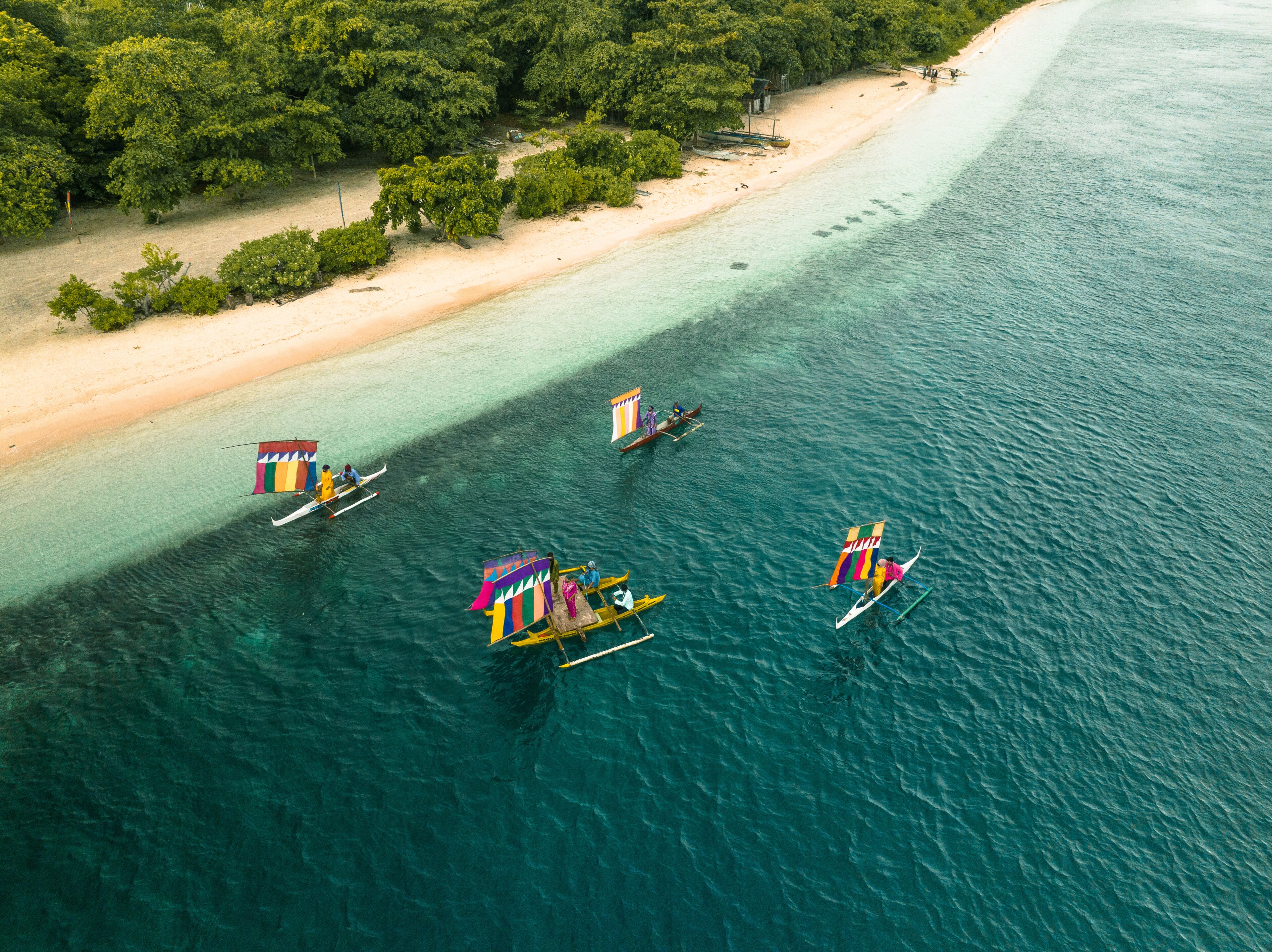 A beach with clear turquoise waters, boats with colorful sails, and dense green foliage in the background.