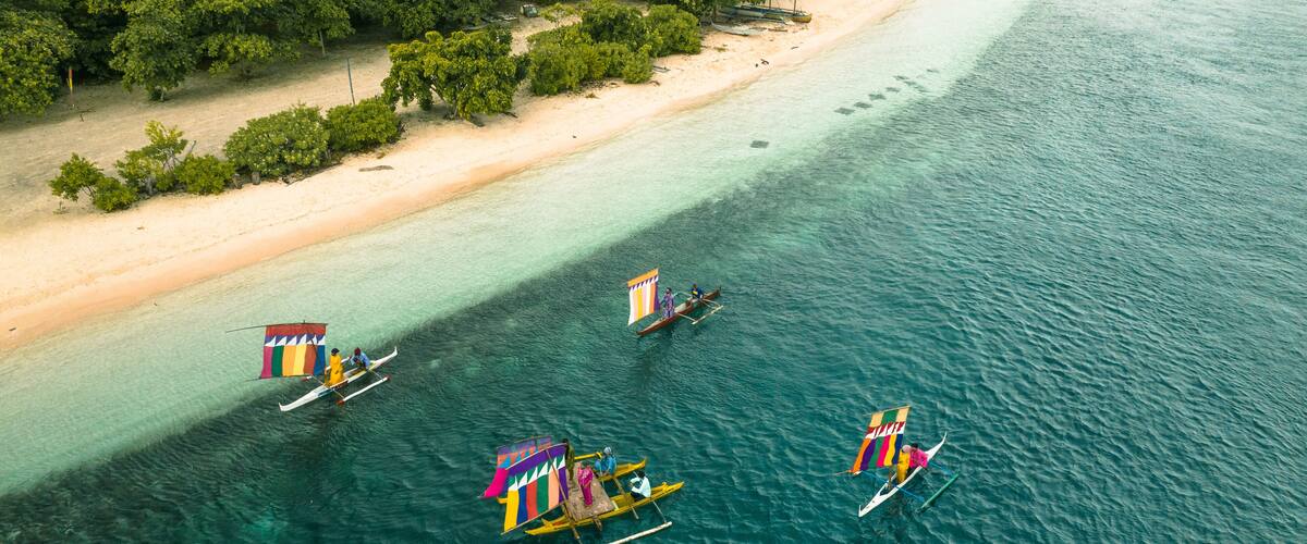 A beach with clear turquoise waters, boats with colorful sails, and dense green foliage in the background.